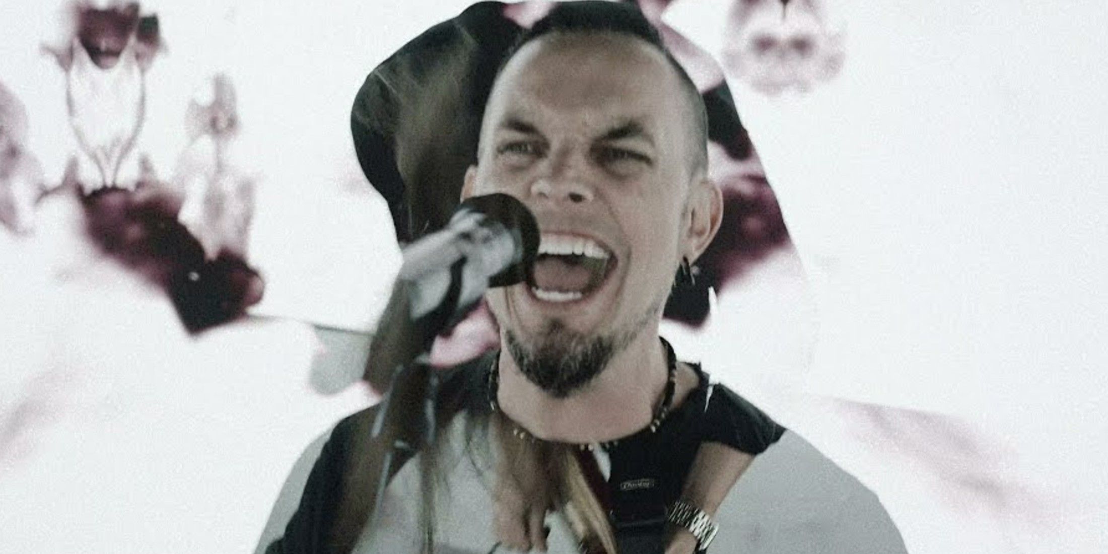 tremonti stands at a microphone with his mouth open to sing. He has a short black beard and short black hair. The background is white with splashes of roses.