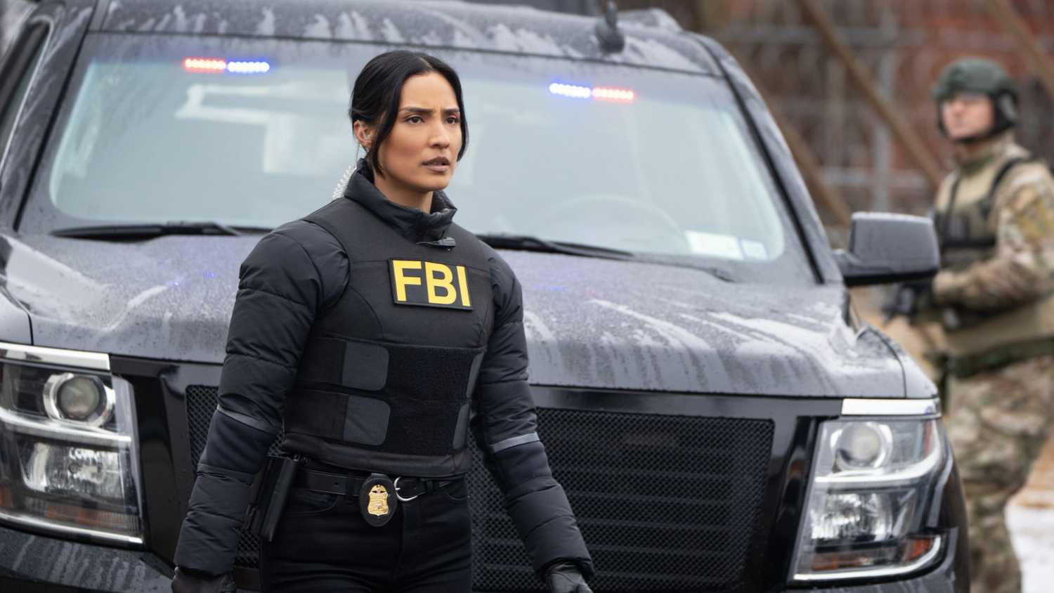 FBI Dani standing in front of a car where a guy in camoflauge is holding a gun