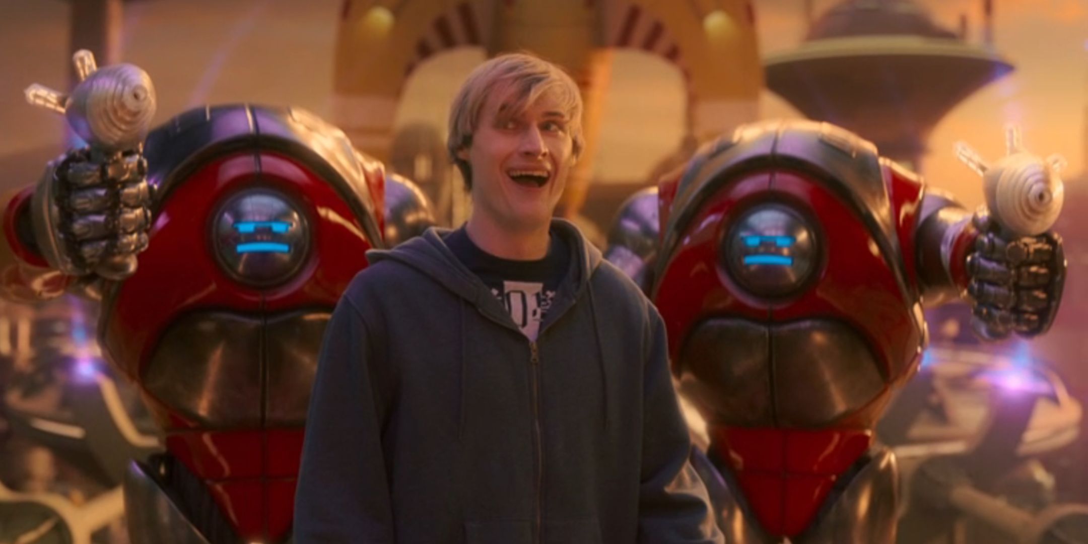 Jonny Green as Alan Budd smiling with Robots behind him in Doctor Who