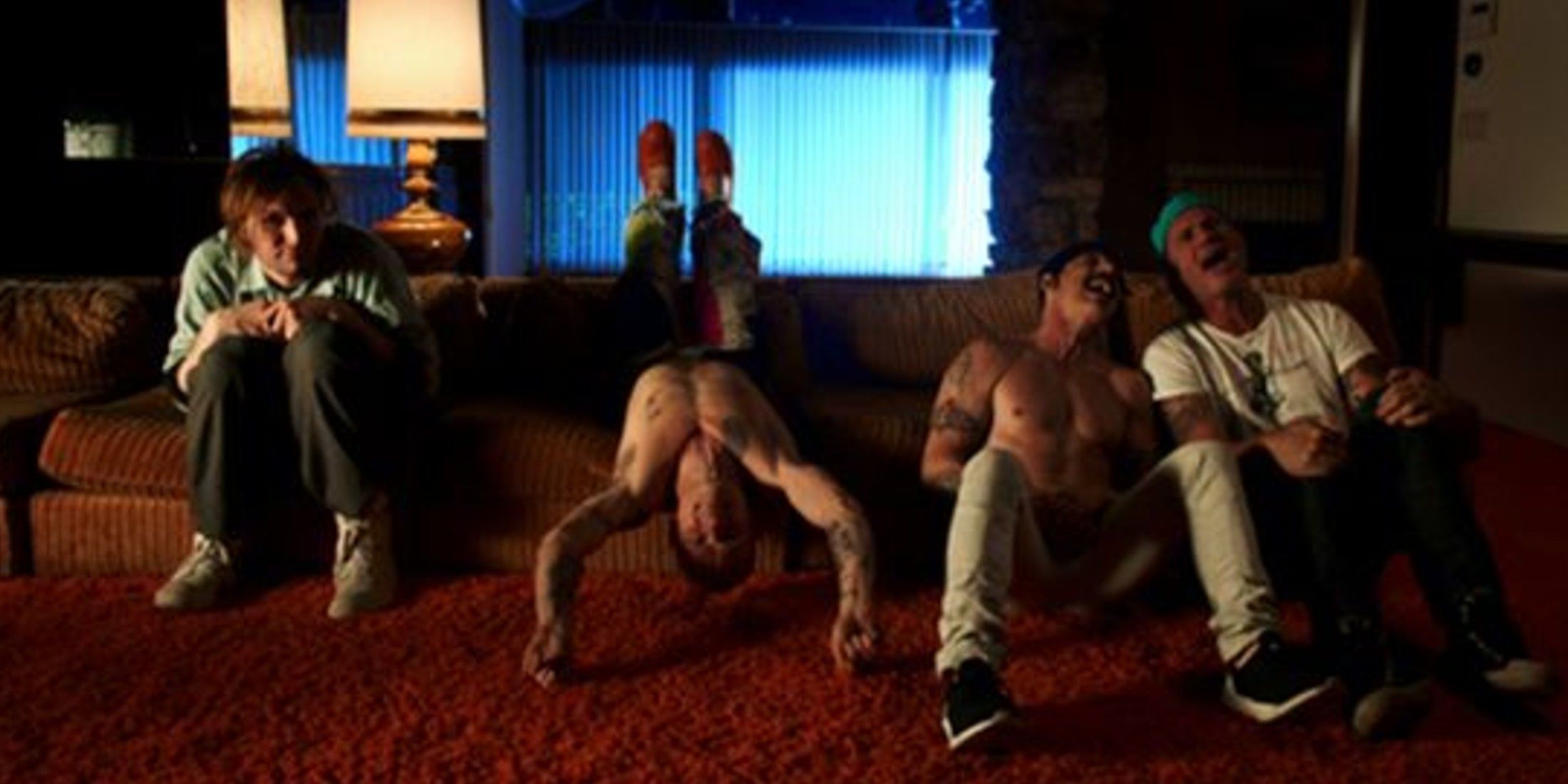 There is a red carpet, and a couch that is a tan color. There are long blinds and two lamps in the background. The person on the left has on a long shirt, jeans, and white sneakers as he hunches over. One is shirtless and lays upside down with his jeans visible. One sits on the ground shirts less in jeans. The one on the far right has on a teal hat, a white shirt and jeans as he sits on the floor. 