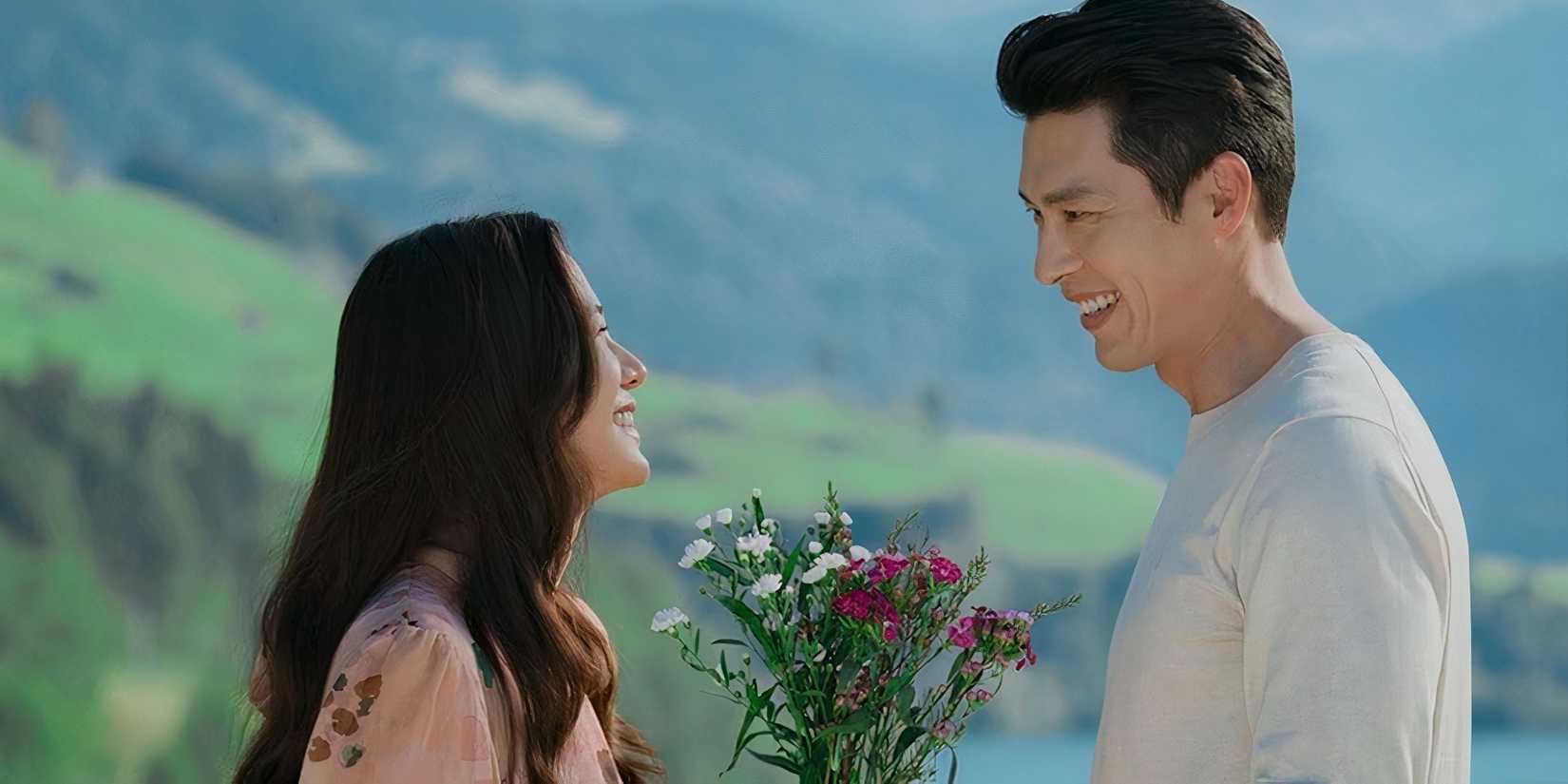 Son Ye-jin and Hyun Bin stand in front of one another smiling with a bouquet of flowers in between them.