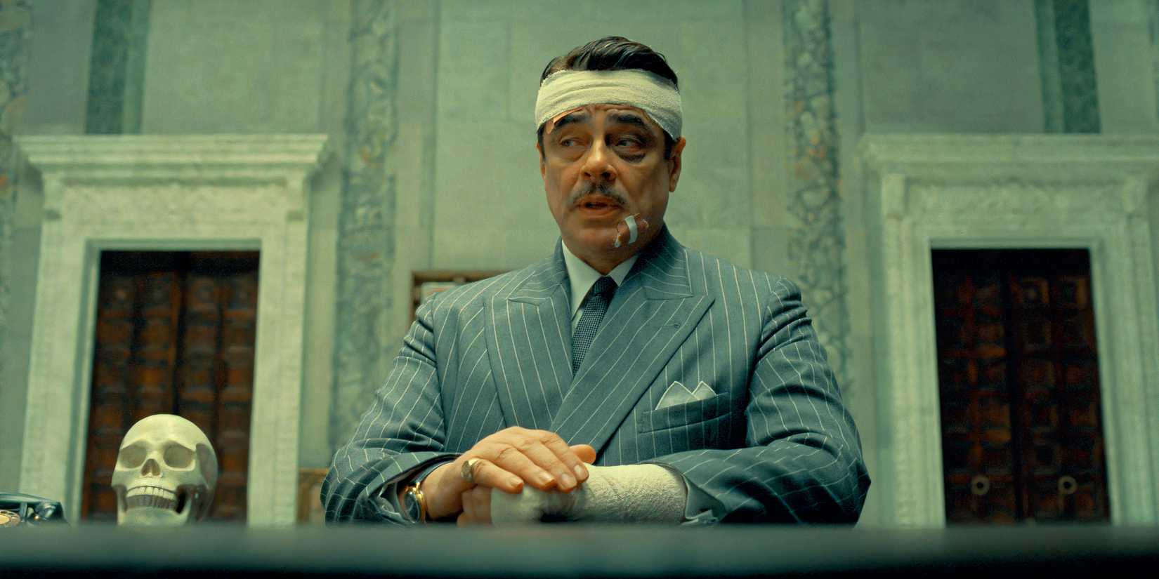 a bandaged benicio del toro sitting with his arms resting on a table in the phoenician scheme