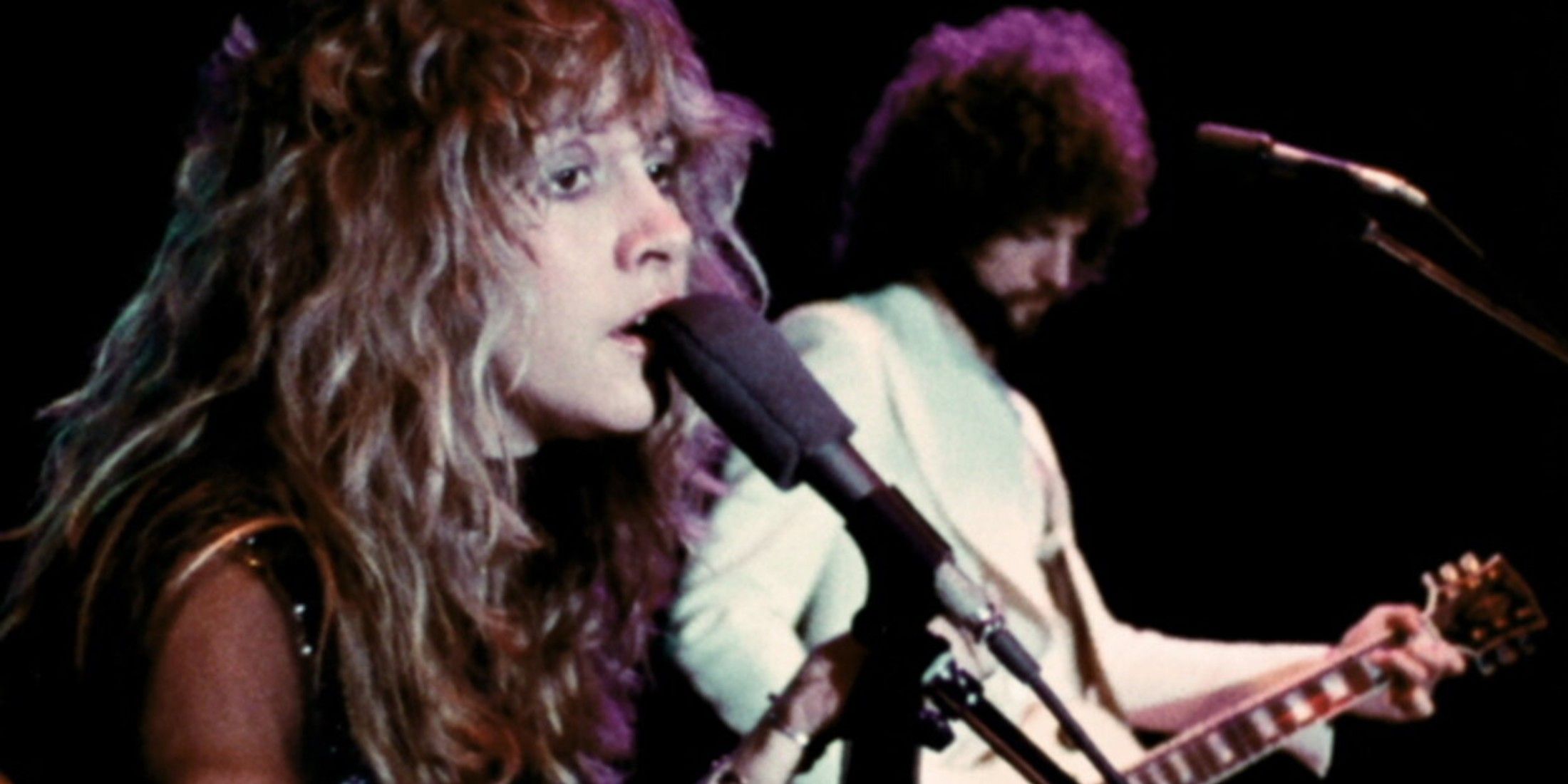 The singer is up close on shown from shoulder up, singing into a microphone. Her hair is long, and her dress is black. The guitarist wears white and plays the guitar diagonal to her. A microphone is in front of him