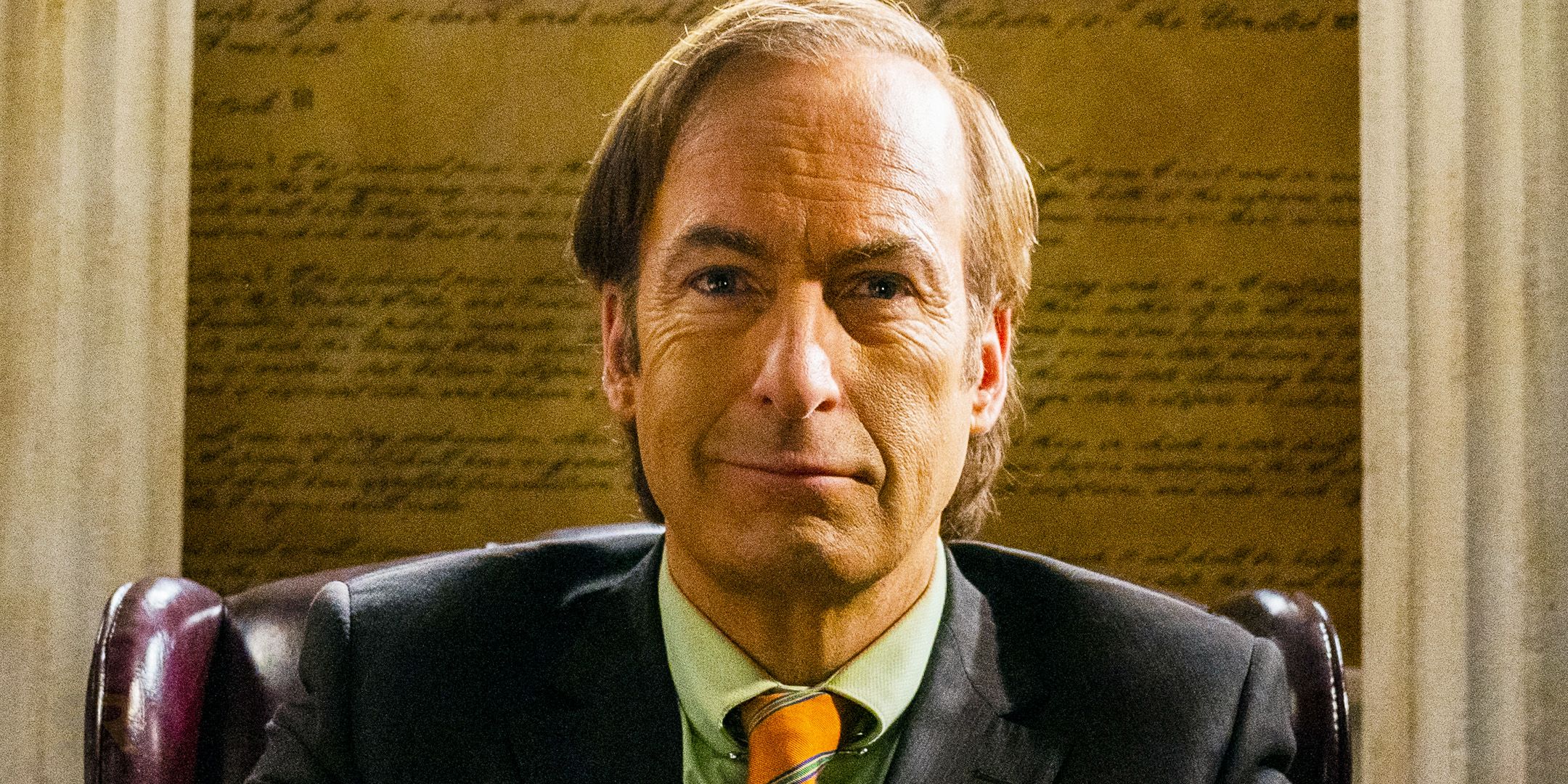 Jimmy McGill/Saul Goodman (Bob Odenkirk) sorrindo em seu escritório na 6ª temporada de Better Call Saul