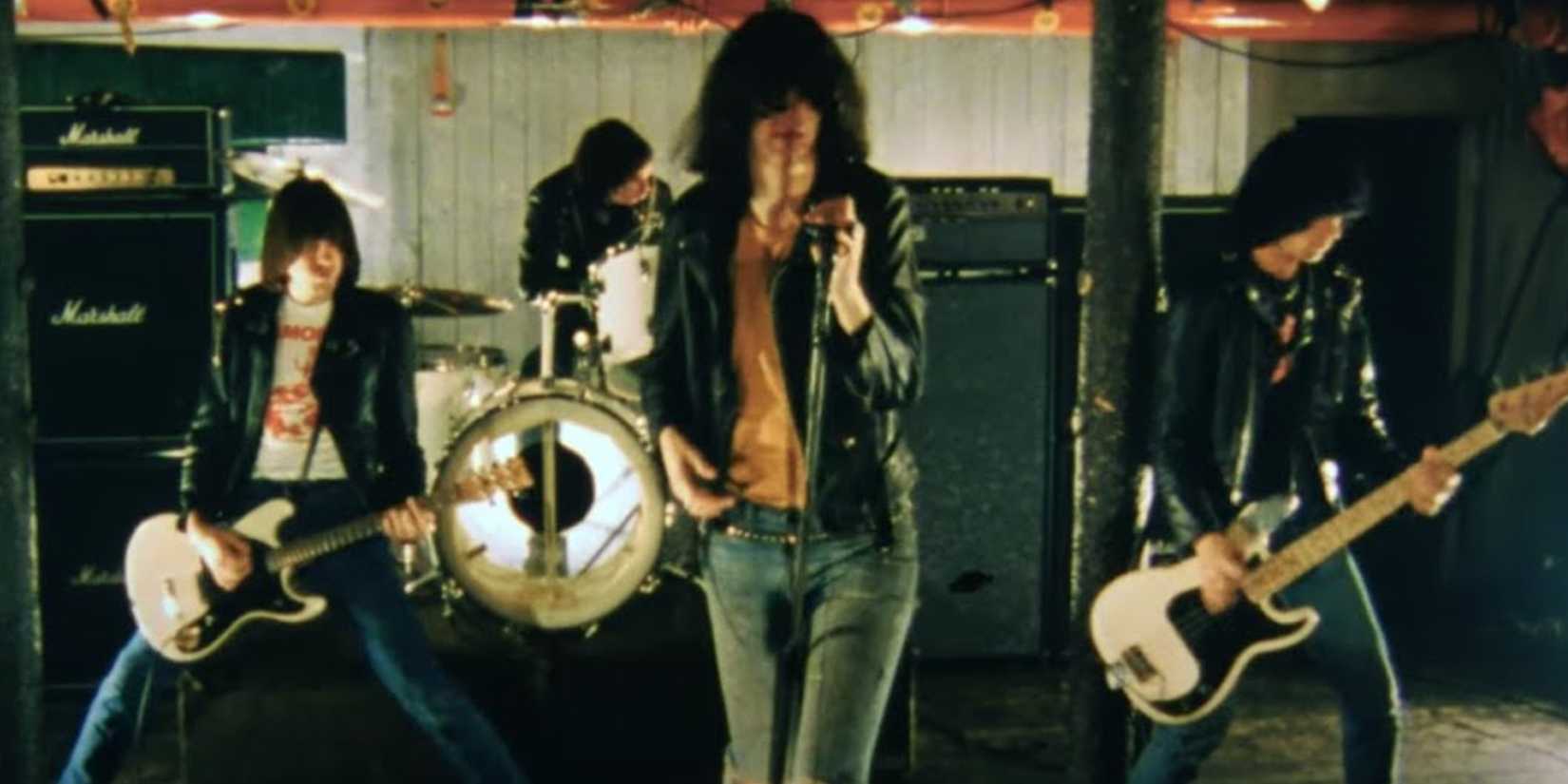 Entourage's 10 Best Fake Movies 6 One plays a white bass guitar on the left. Behind him is a drummer on a white drum kit. The singer is shirtless, but wears a leather jacket as he holds a chord microphone and sings. The one on the right plays the guitar. Each member has on leather jackets.