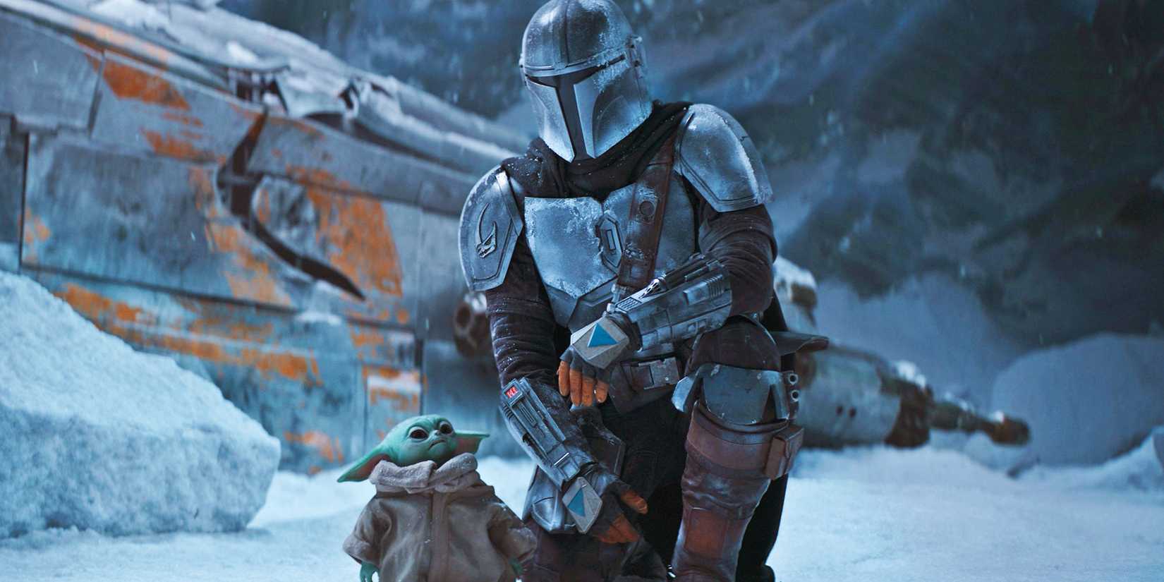 Din Djarin (Pedro Pascal) kneels beside Grogu in the snow on Maldo Kreis in front of the wrecker Razor Crest.