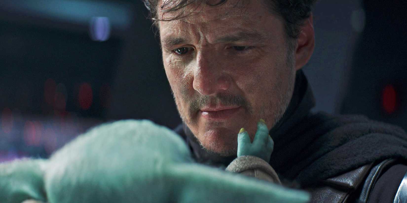 Din Djarin (Pedro Pascal), without his helmet, smiles down at Grogu in his arms, who's holding his face.