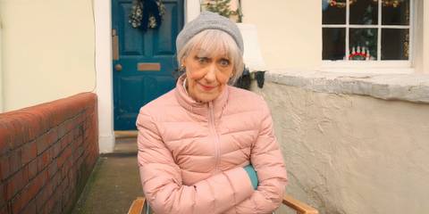 Anita Dobson as Mrs. Flood giving a dubious look while sitting in a chair with arms crossed
