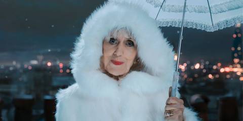 Anita Dobson as Mrs. Flood giving a tight-lipped smile wearing a white hooded coat and holding an umbrella