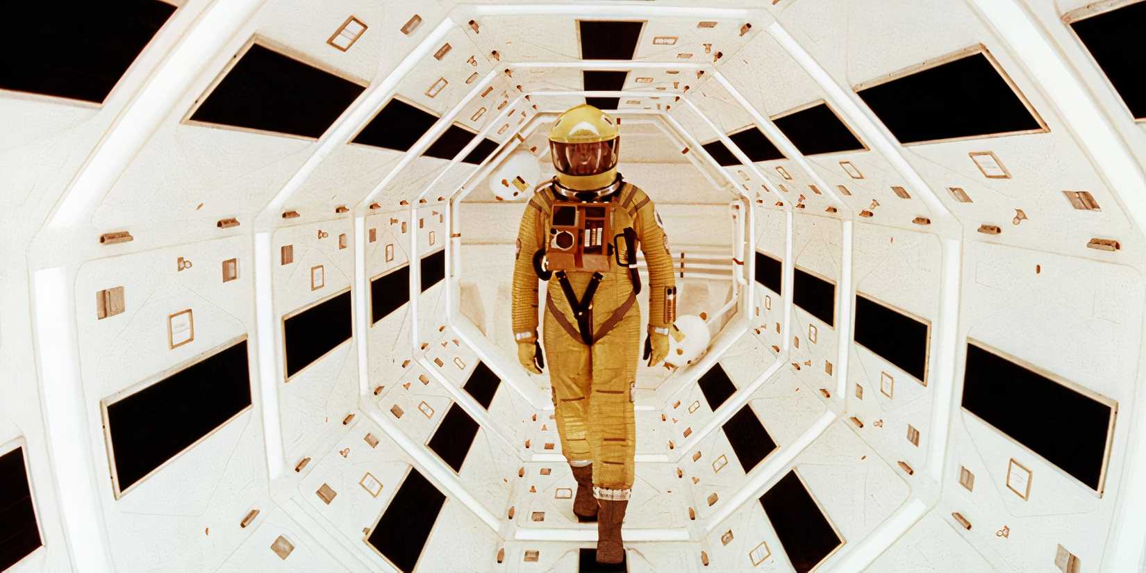 An astronaut in a yellow space suit walks through a long white hallway in 2001 A Space Odyssey