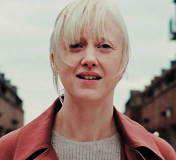 A woman in a red coat stands between two buildings in Possessor.