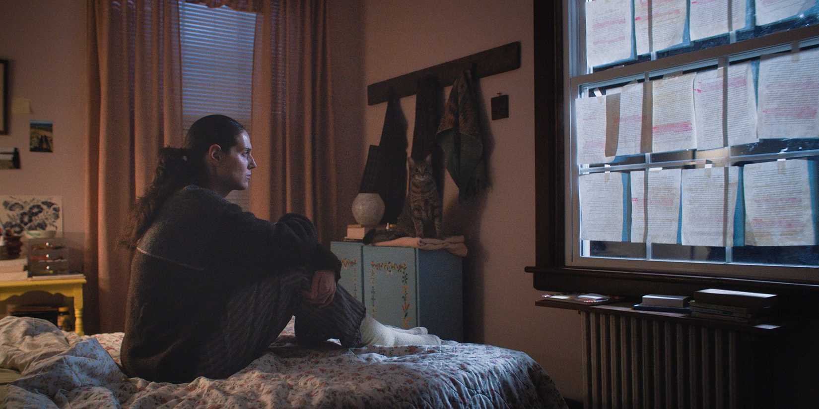 Agnes sentada em sua cama olhando para a janela, coberta de papéis com sua tese, em Sorry, Baby