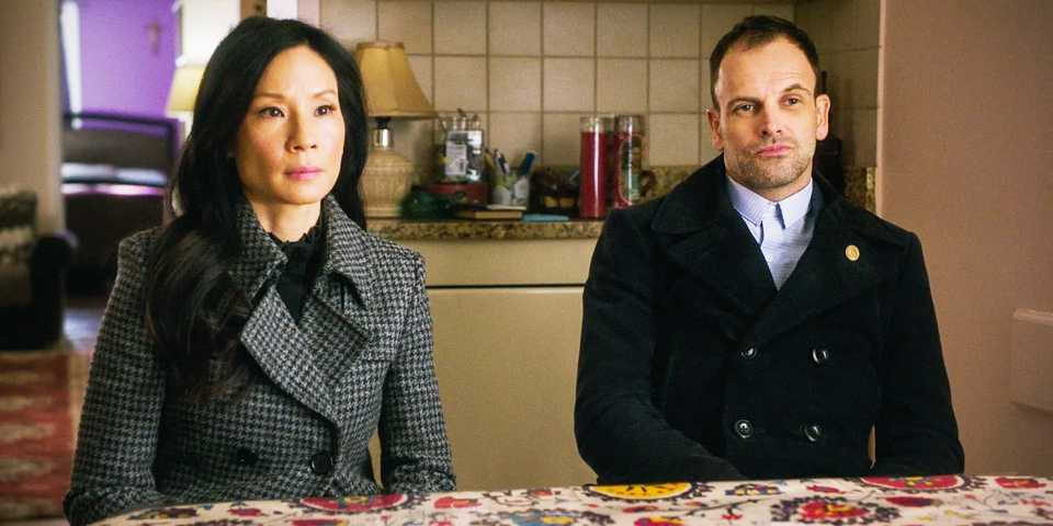 Elementary Joan Watson (Lucy Liu) and Sherlock Holmes (Jonny Lee Miller) sitting next to one another.