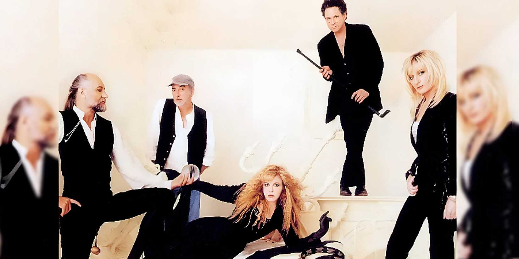 The background is a white back drop. On the far left stands someone in black pants, a black vest, and a white undershirt. He holds the hand of girl laying on her side in a black dress. Behind her stands someone in blue jeans, a white under shirt, a black vest, and a grey hat. Next to him is someone standing with their legs crossed. They have on black pants, a black coat, and holds a black cane. In the far right hand corner, a girl stands in black pants, a black coat with a white under shirt, and