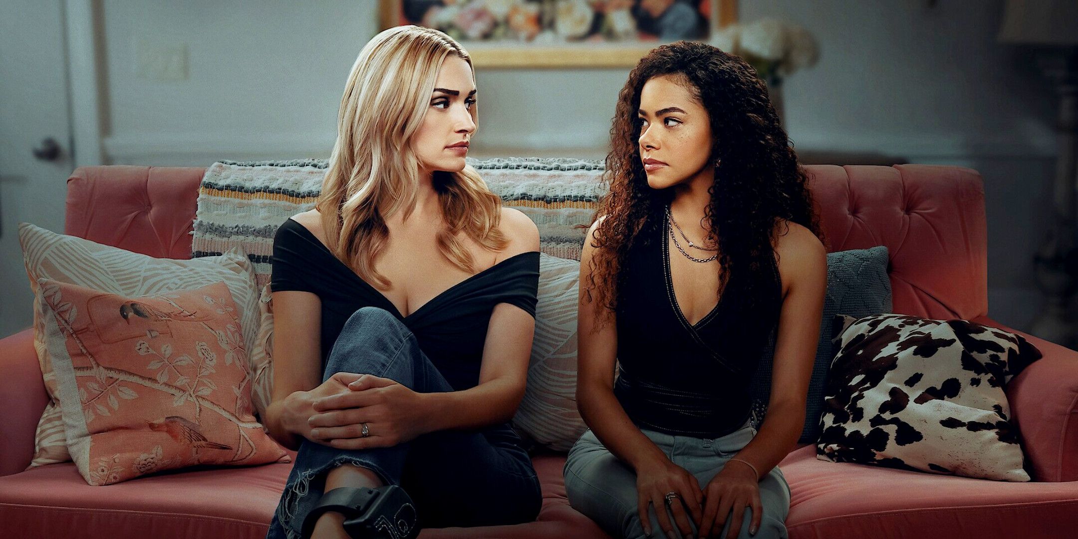 Brianna Howey as Georgia Miller and Antonia Gentry as Ginny Miller in Ginny & Georgia season 3. They're sitting on a pink sofa looking at each other, wearing jeans and black tops.