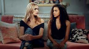 Brianna Howey as Georgia Miller and Antonia Gentry as Ginny Miller in Ginny & Georgia season 3. They're sitting on a pink sofa looking at each other, wearing jeans and black tops.