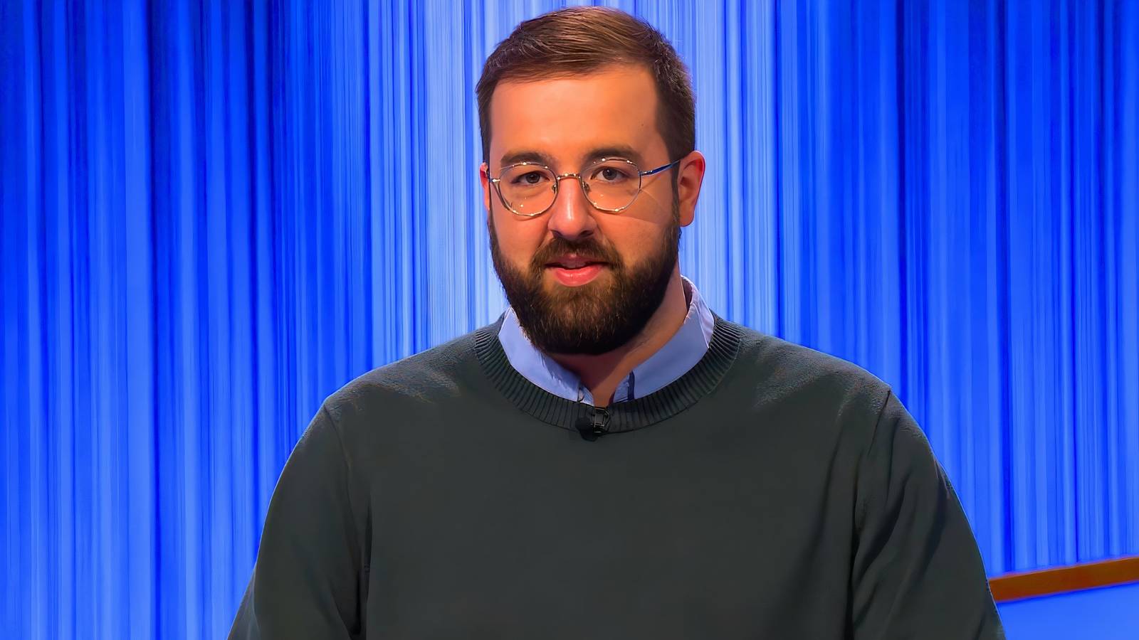 Jeopardy star Andrew Hayes looking serious in a shirt and sweater with a blue background