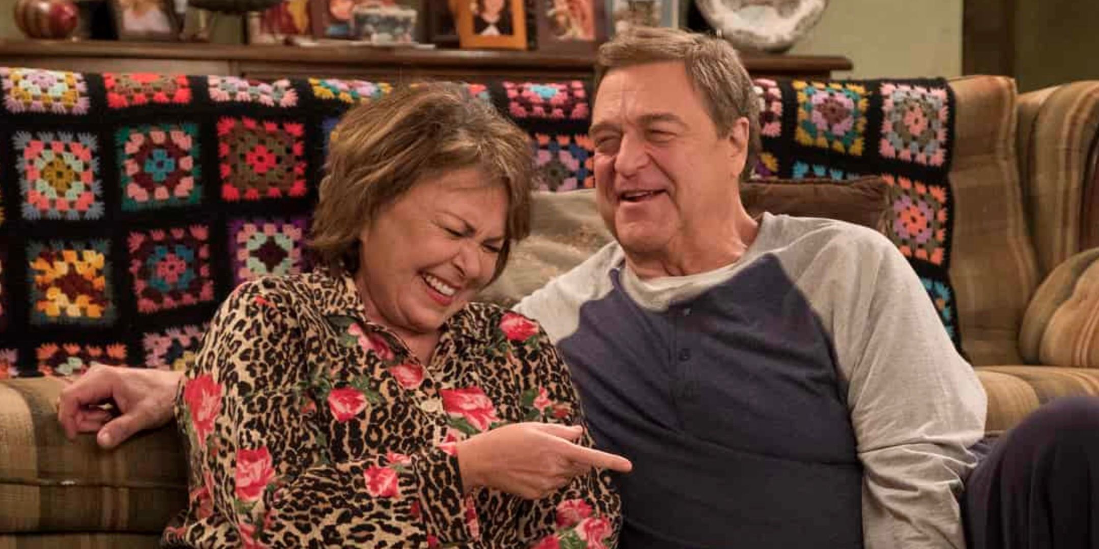 Roseanne Barr's Roseanne and John Goodman's Dan sit beside a couch laughing in Roseanne season 10
