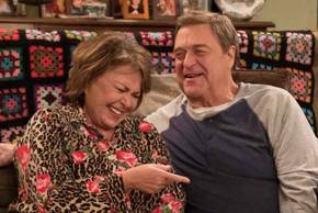 Roseanne Barr's Roseanne and John Goodman's Dan sit beside a couch laughing in Roseanne season 10