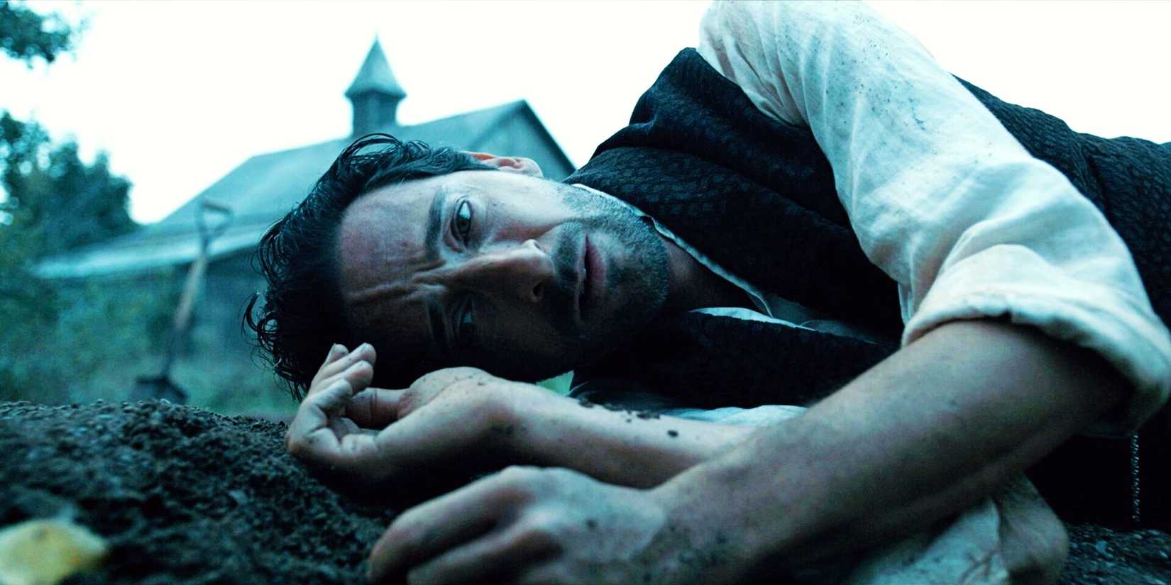 Adrien Brody laying down on a grave in Chapelwaite with a church behind him