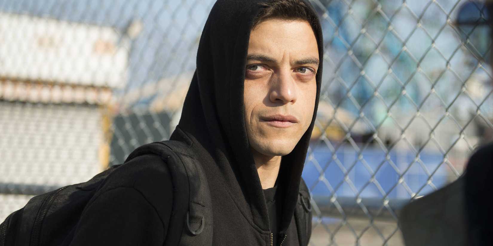 Elliot com seu capuz preto levantado ao lado de uma cerca de arame em Mr. Robot