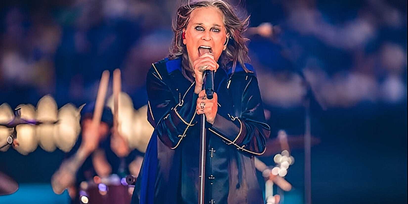 The background behind Ozzy Osbourne is blurry, but a drummer is somewhat visible. Ozzy is shown from the waist up, wearing a long coat and singing into a microphone, as his long hair flows.