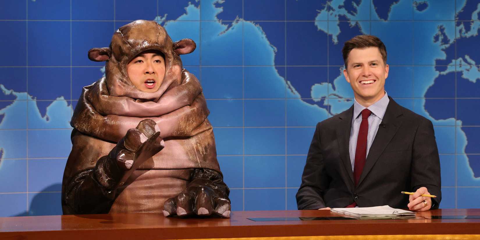 Colin Jost smiling while sitting next to a hippo on Saturday Night Live's Weekend Update