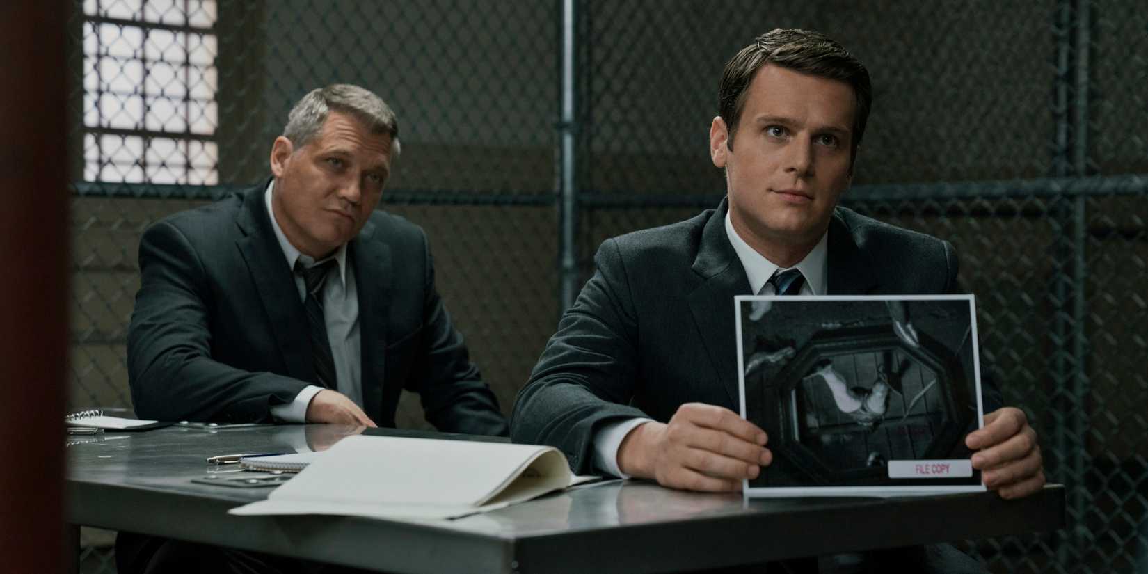 Holden Ford and Bill Tentch holding up a picture in a prison, while interviewing someone who is off-screen, in Mindhunter.