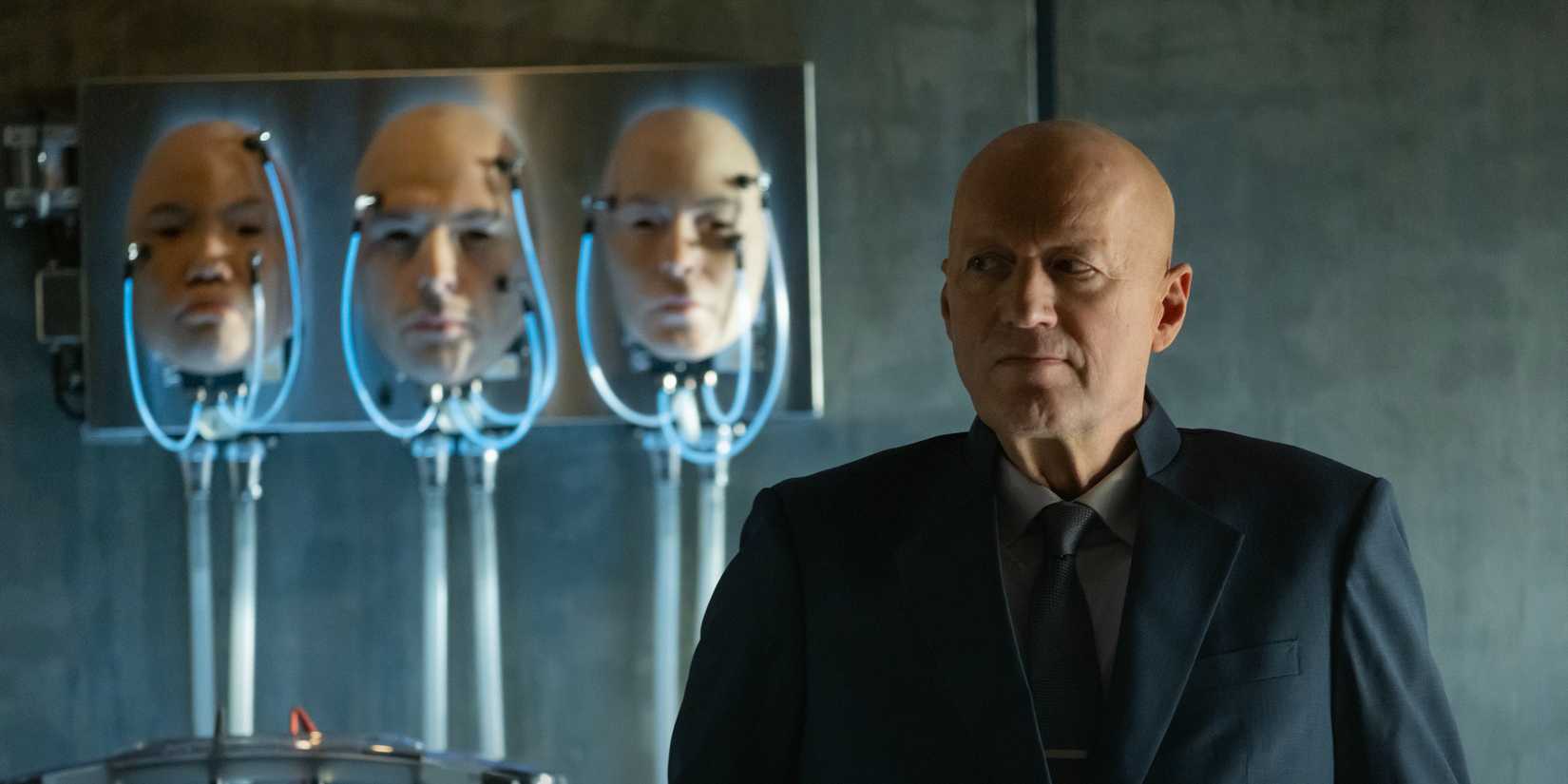 Ade Edmondson like Atom Eins. Standing next to a few prosthetic heads in Alien: Earth Episode 4