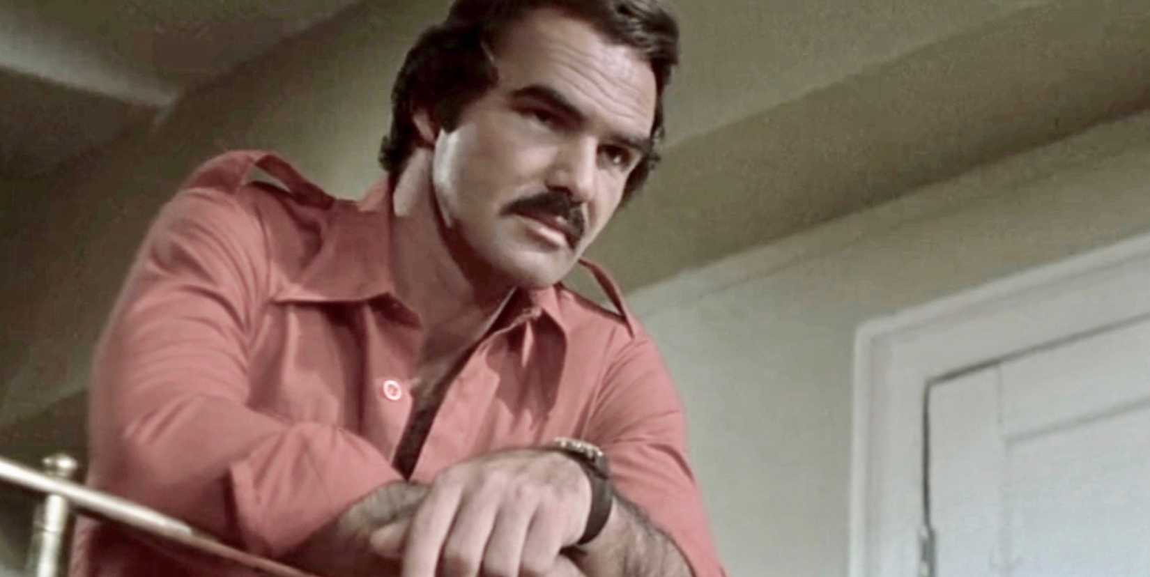 Burt Reynolds leans against a bed frame and looks angry in Gator