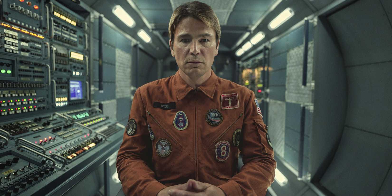 Josh Hartnett seated, hands folded in an orange combination in black mirror