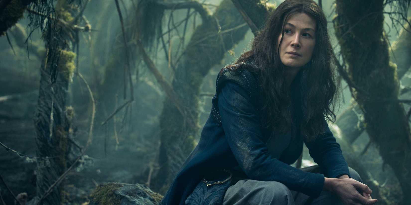 Rosamund Pike as Moiraine Damodred, sitting on a rock within a hazy forest, in The Wheel of Time