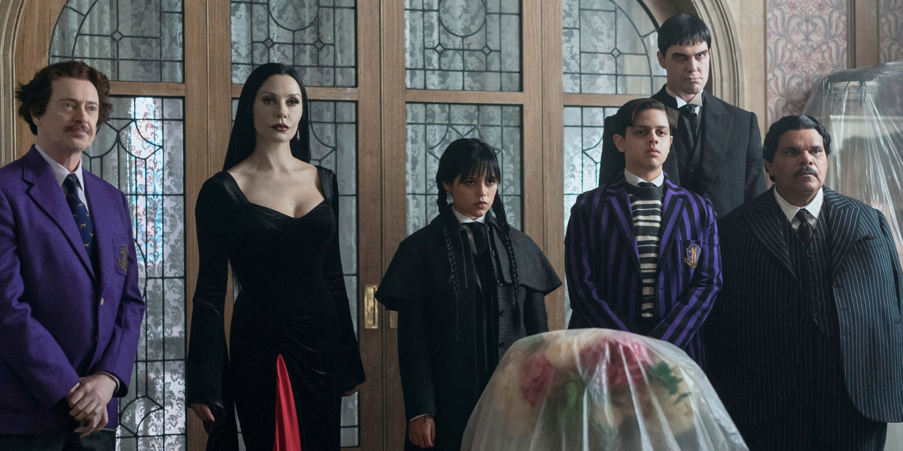 Barry Dort (Steve Buscemi), Morticia (Catherine Zeta-Jones), Wednesday (Jenna Ortega), Pugsley (Isaac Ordonez), Lurch (Joonas Suotamo), and Gomez Addams (Luis Guzman) standing in front of a door and looking forward in Wednesday season 2