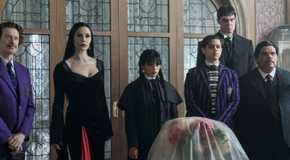 Barry Dort (Steve Buscemi), Morticia (Catherine Zeta-Jones), Wednesday (Jenna Ortega), Pugsley (Isaac Ordonez), Lurch (Joonas Suotamo), and Gomez Addams (Luis Guzman) standing in front of a door and looking forward in Wednesday season 2