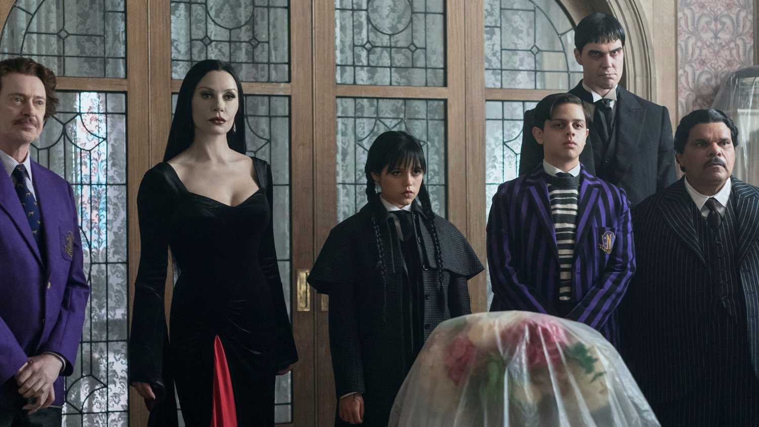 Barry Dort (Steve Buscemi), Morticia (Catherine Zeta-Jones), Wednesday (Jenna Ortega), Pugsley (Isaac Ordonez), Lurch (Joonas Suotamo), and Gomez Addams (Luis Guzman) standing in front of a door and looking forward in Wednesday season 2