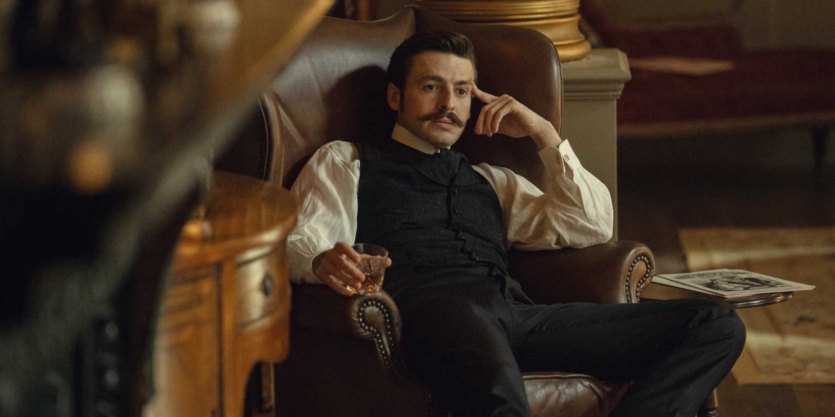 Anthony Boyle as Arthur Guinness sitting on a chair and holding a glass of whiskey in House of Guinness