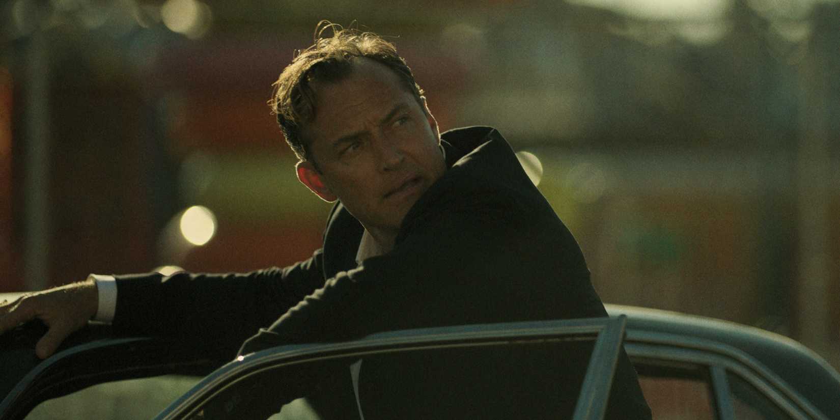 Jake looks over his shoulder while leaning on a car in Black Rabbit