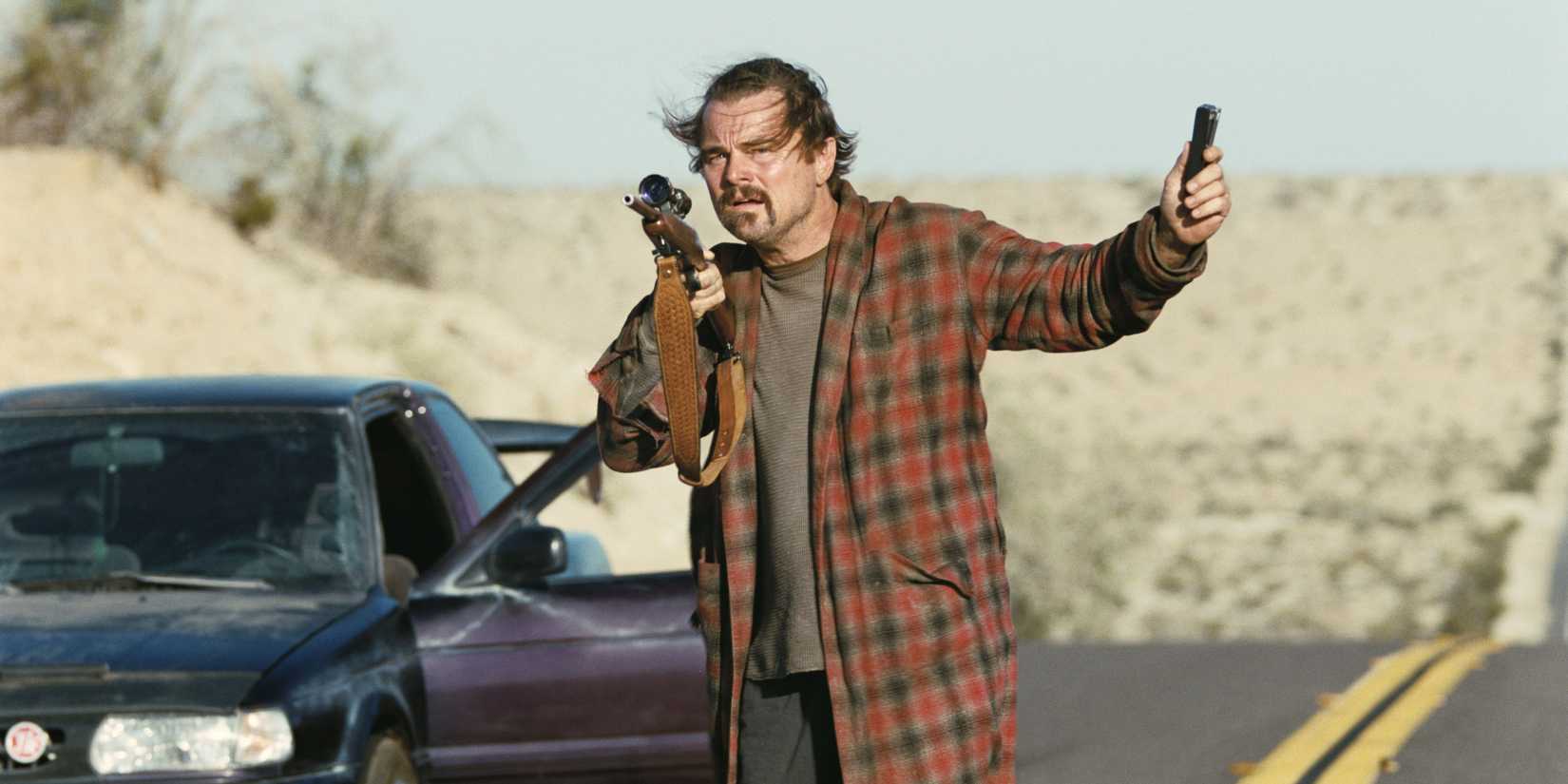 Bob holding a rifle in one hand and the authenticator in another in One Battle After Another