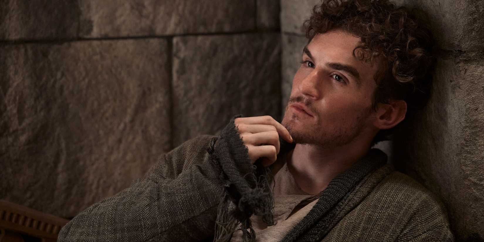 Dónal Finn as Mat Cauthorn lying against a stone wall scratching his chin in The Wheel of Time