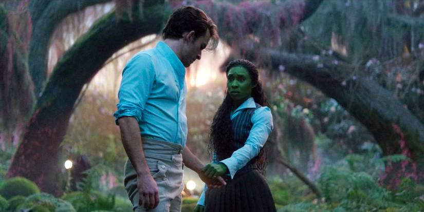 Fiyero stands half-turned toward Elphaba, holding her hand, in the forest in Wicked