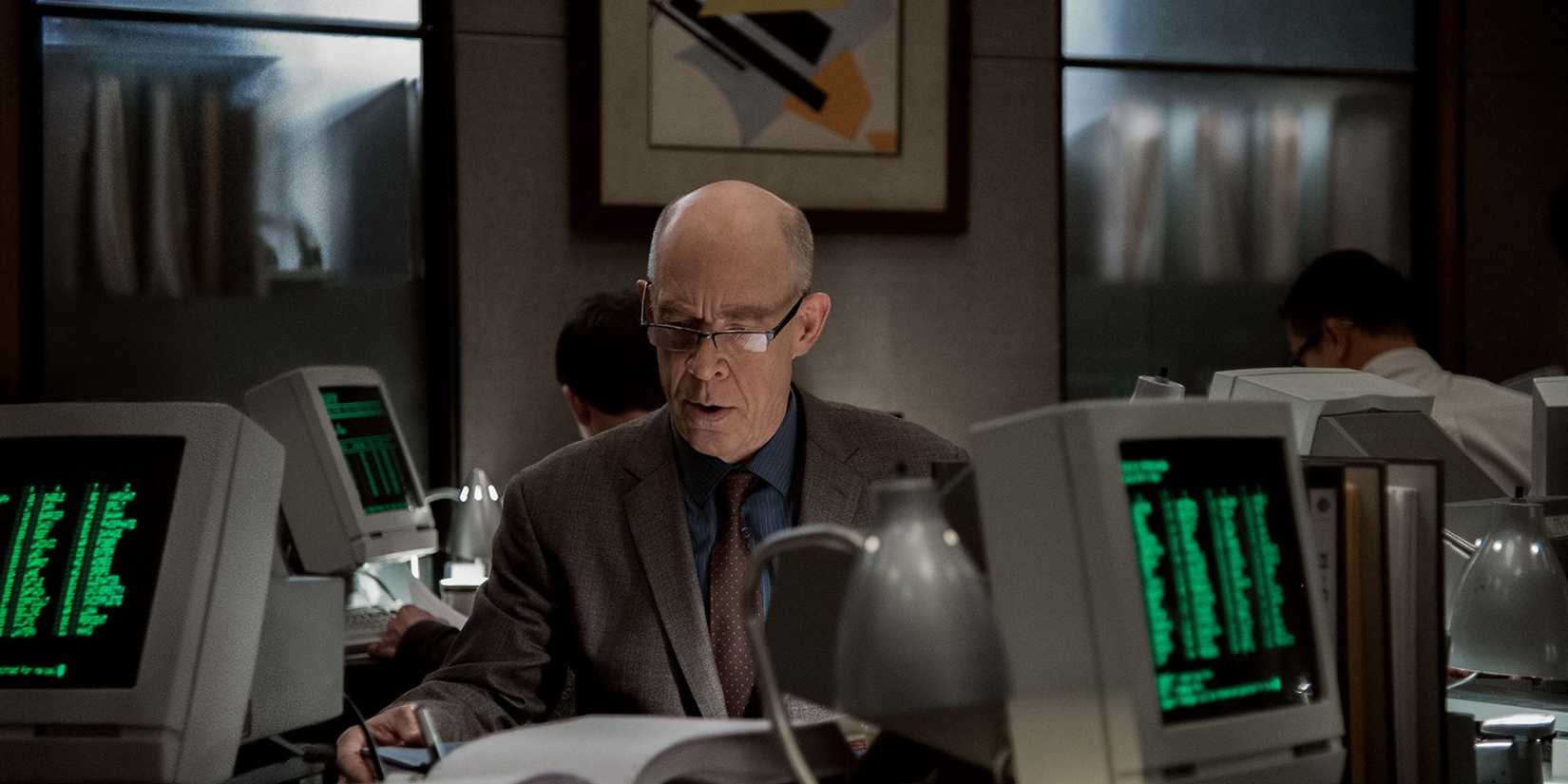 JK Simmons como Howard Silk na contraparte, trabalhando em uma mesa de escritório com computadores antigos ao seu redor