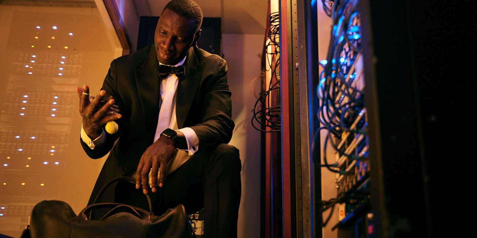 Omar Sy as Assane Diop dressed in a tux, investigating something in his hand next to a data server in Lupin