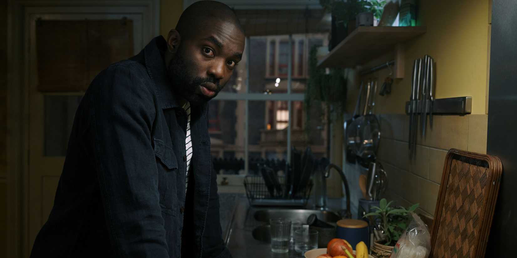 PAAPA Essiedu como George Addo no projeto Lazarus, encostado a um balcão de cozinha