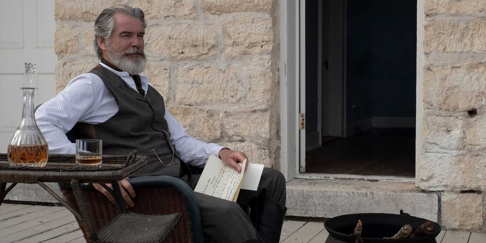 Pierce Brosnan as Eli McCullough lounging on an old Western porch with a letter in hand, and some scotch , in The Son