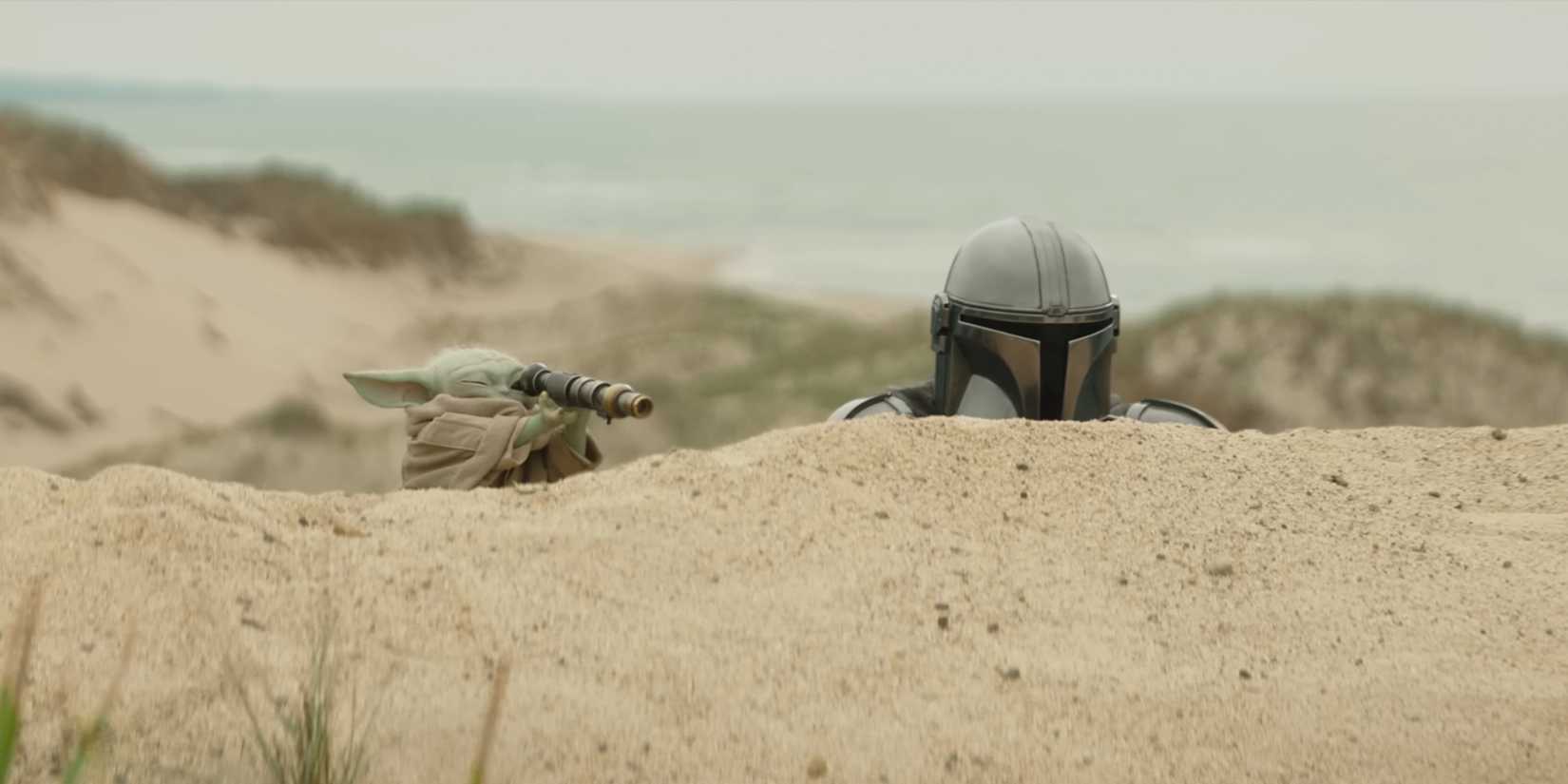 Din Djarin and Grogu peek their heads up from behind a sand dune as Grogu looks through a scope.
