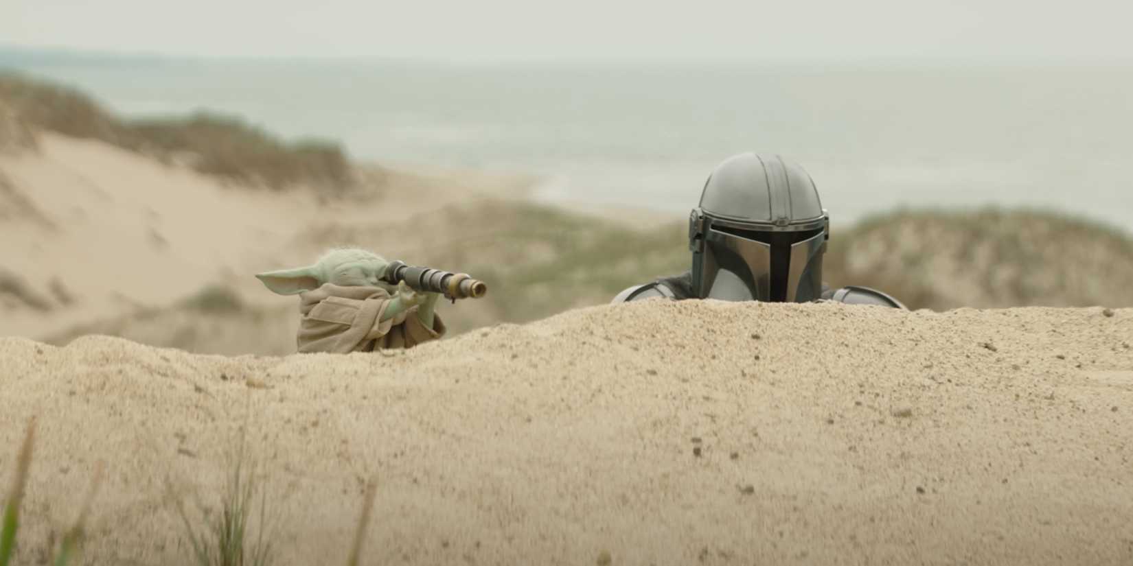 Din Djarin and Grogu peek their heads out from behind a sand dune as Grogu looks through a scope.