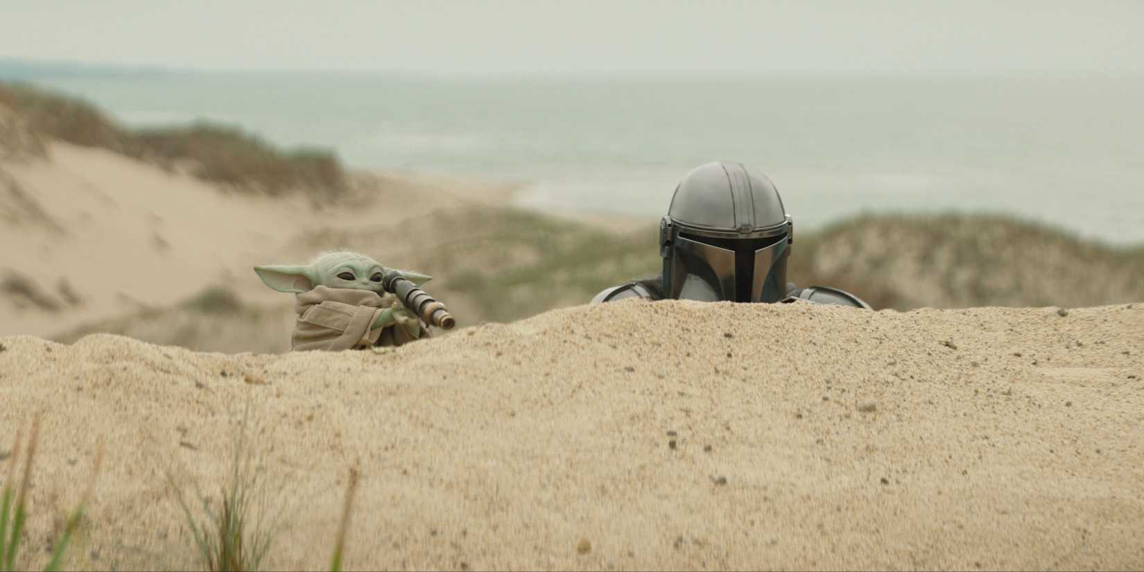Grogu and Mando hiding behind a sand dune in The Mandalorian & Grogu