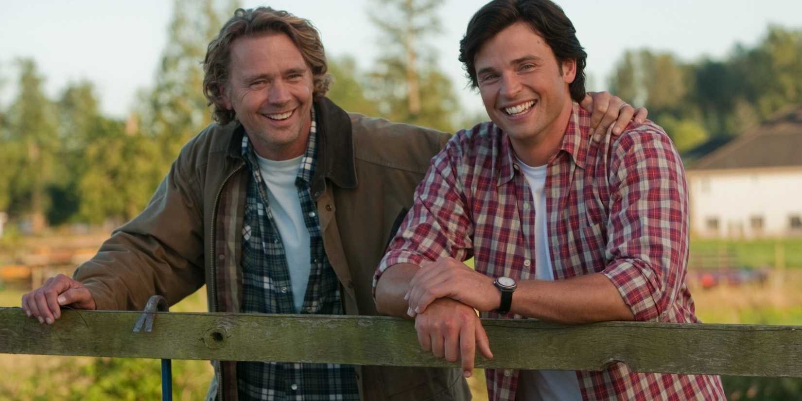 Tom Welling y John Schneider se apoyan en un cercado de una granja riendo juntos en Smallville