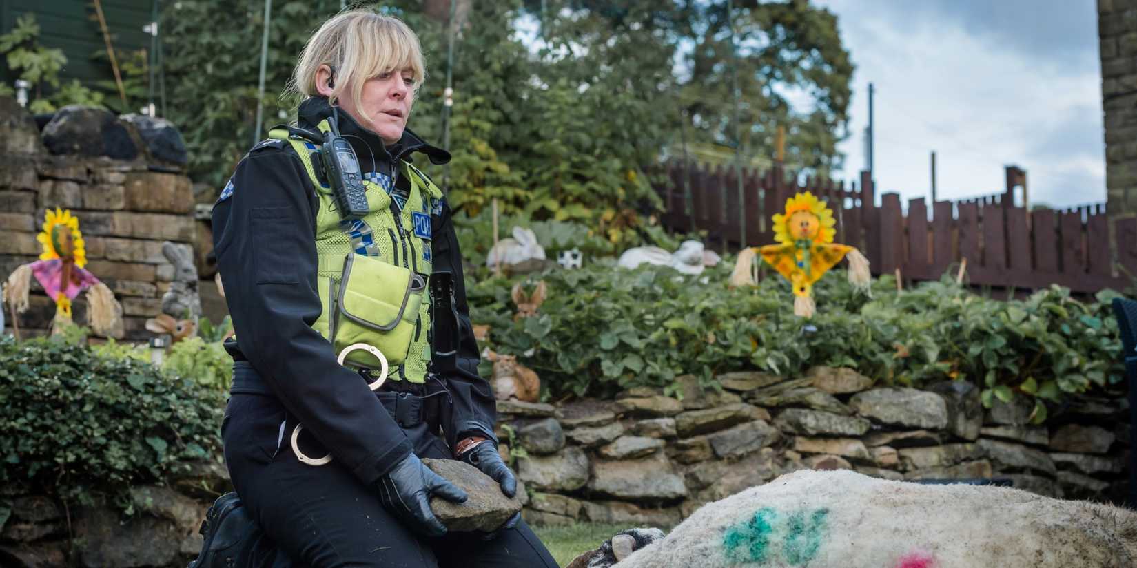 Sarah Lancashire as Sgt Catherine Cawood, kneeling in a garden and looking exhausted, while holding a rock next to a sheep in Happy Valley.