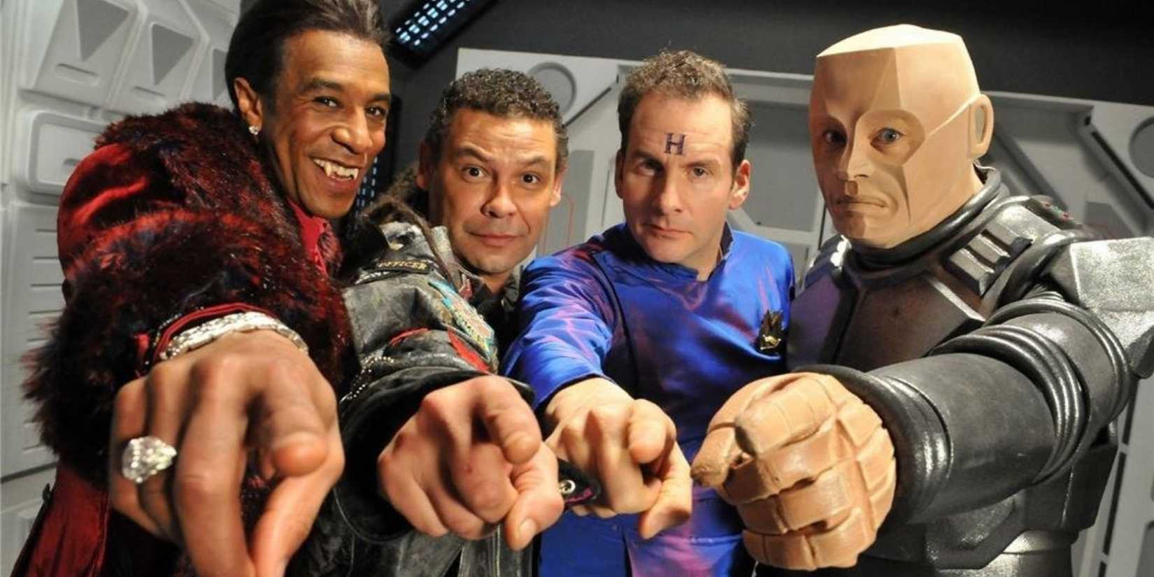 Lister, Rimmer, Kryten, and Cat pointing and smiling on their ship as they pose in Red Dwarf.